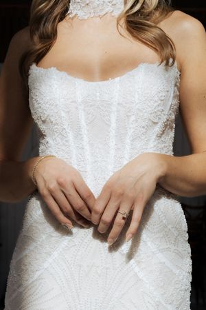 Close-up of a bride in a strapless beaded white wedding gown, hands folded, wearing an engagement ring and gold bracelet.