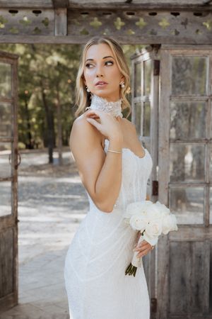 Bride in white beaded gown holding bouquet of white roses poses outdoors near rustic wooden doors.