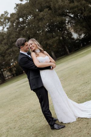 Bride and groom sharing romantic embrace on green lawn, bride in lace gown, groom in black tuxedo.