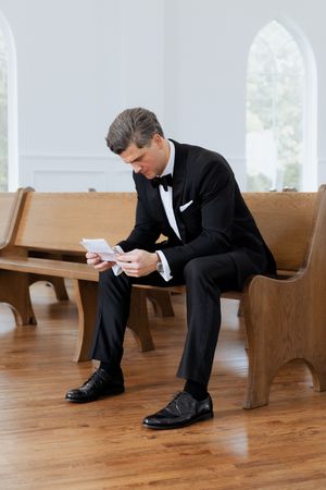 Groom in a classic black tuxedo sits on a church pew reading notes before his wedding ceremony.