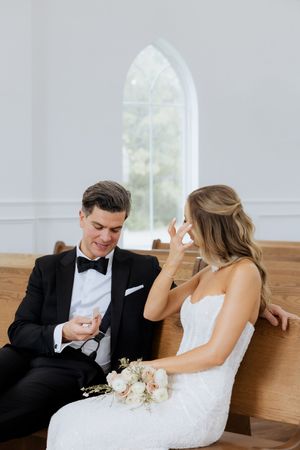 Bride and groom sitting on a church pew, sharing a tender moment in a bright white chapel with arched windows.