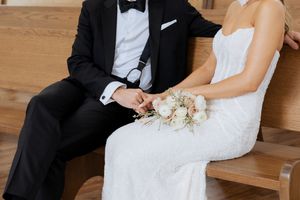 Bride and groom holding hands on wooden church pew, bride holding white floral bouquet.