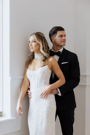 Elegant bride in white lace gown and pearl choker stands with groom in black tuxedo against white wall.