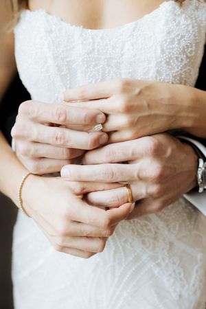 Close-up of newlyweds hands intertwined showing wedding rings, bride in white lace dress.