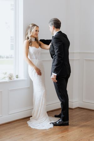 Bride in white lace gown and groom in black tuxedo share an intimate first look moment by a bright window.