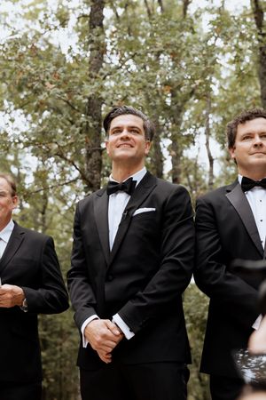 Smiling groom in black tuxedo with bow tie stands with groomsmen during outdoor wedding ceremony.