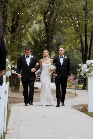 Bride in white gown walks down outdoor aisle flanked by two men in tuxedos.