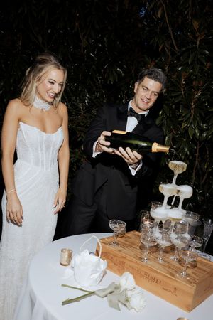 Bride and groom in formal attire pour champagne tower at outdoor wedding reception at night.