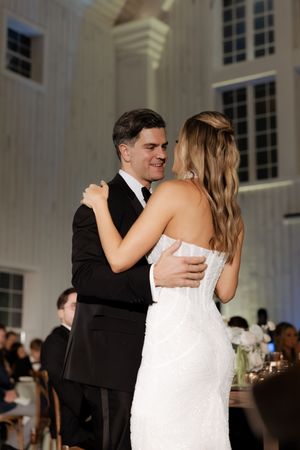 Bride and groom share their first dance, smiling warmly in a beautifully lit white wedding reception venue.