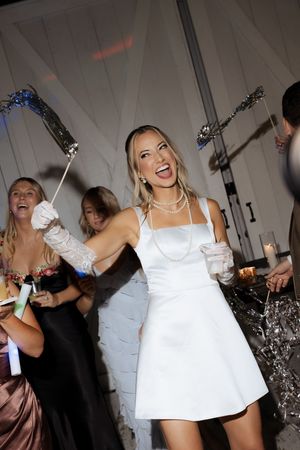 Joyful bride in white mini dress with pearl necklace and gloves dancing at wedding reception.