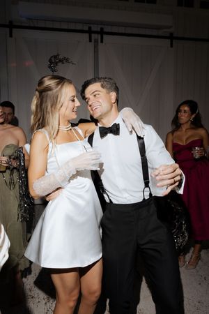 Bride in short white dress dances with groom in tuxedo at wedding reception.