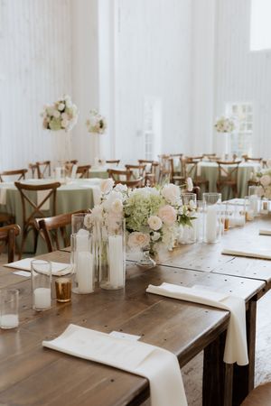 Elegant wedding reception venue with wooden farm tables, white floral centerpieces, candles, and sage green accent tables.