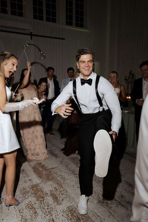 Groom in suspenders and bow tie dances energetically with guests celebrating at a lively wedding reception.