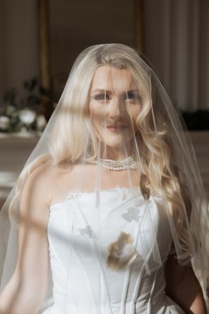A bride in a white wedding dress and veil poses in an elegant room with crystal chandeliers.