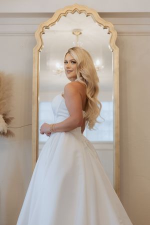 A bride in a flowing white ballgown poses elegantly against an ornate gold-framed mirror.