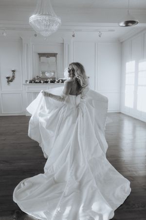 A flowing white gown twirls gracefully across a hardwood floor beneath a crystal chandelier in a bright studio.