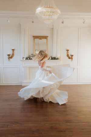 A flowing white wedding dress twirls across a hardwood floor in an elegant room with chandeliers.