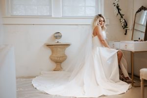 A bride in a flowing white wedding gown with a long train poses in an elegant white room with ornate decor.