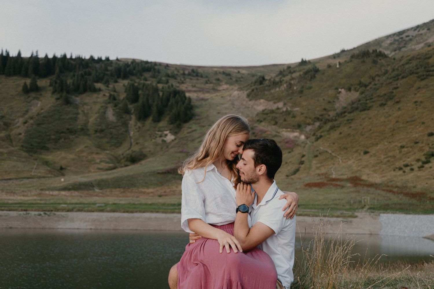 A couple embraces against a scenic mountain backdrop with a lake and evergreen trees.