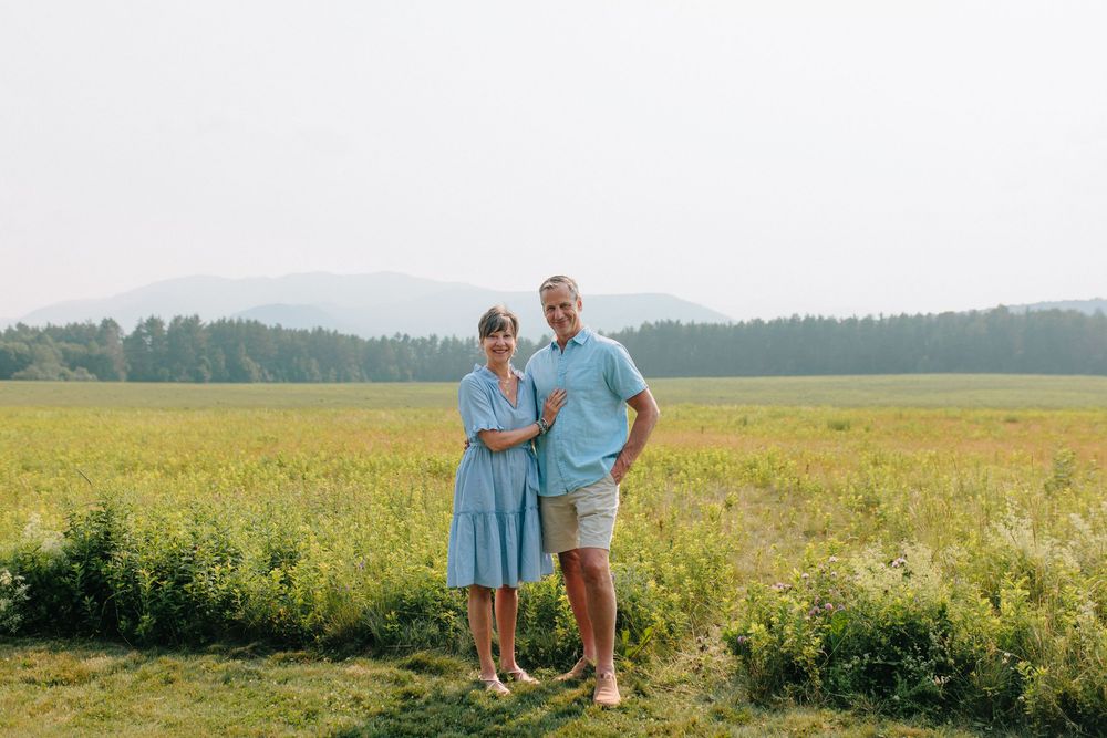 Couple in light blue and white outfits stands together in flowering meadow with forest backdrop.