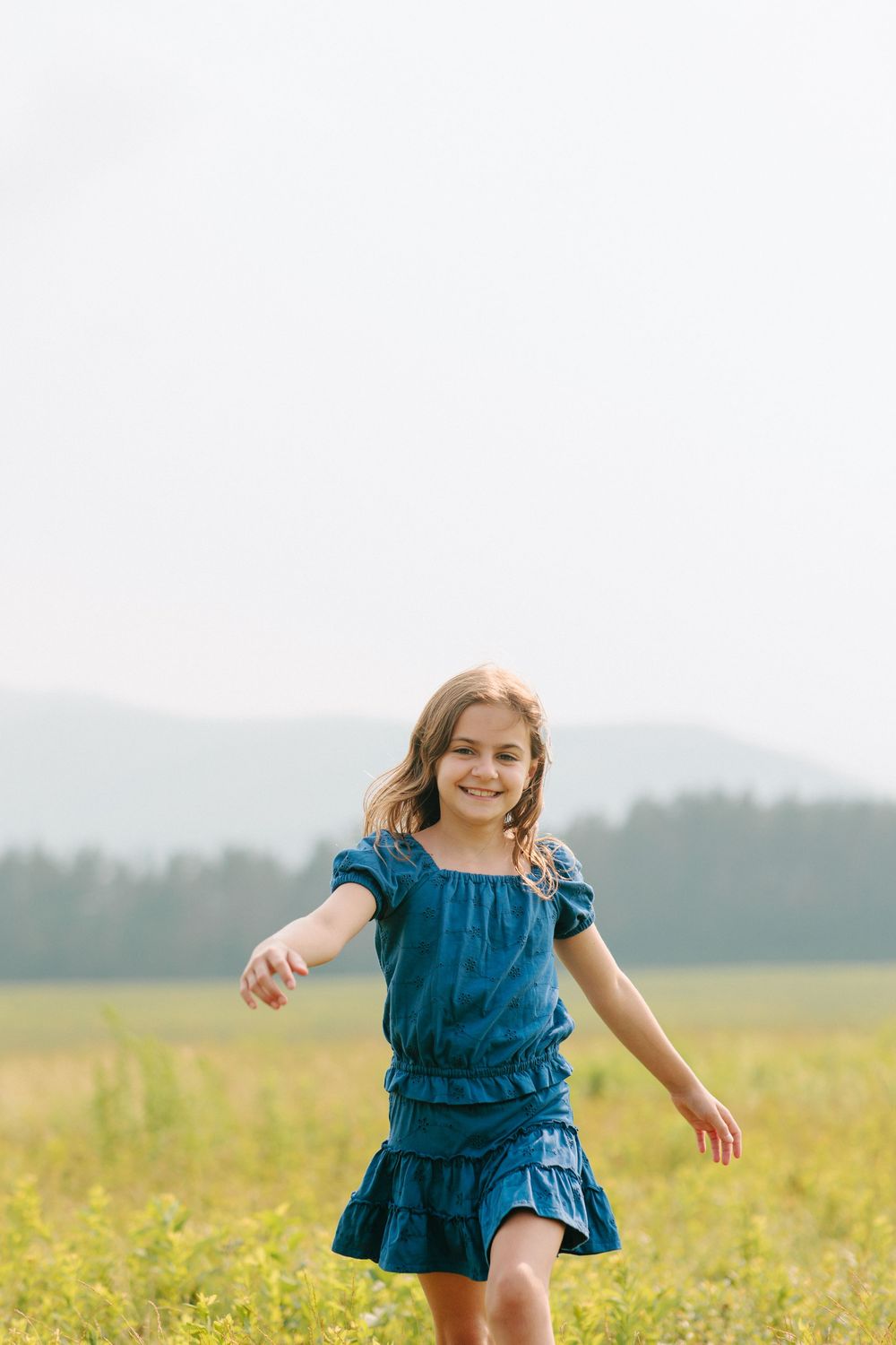 Young girl in blue dress twirls happily in a summer meadow with mountains in the distance.