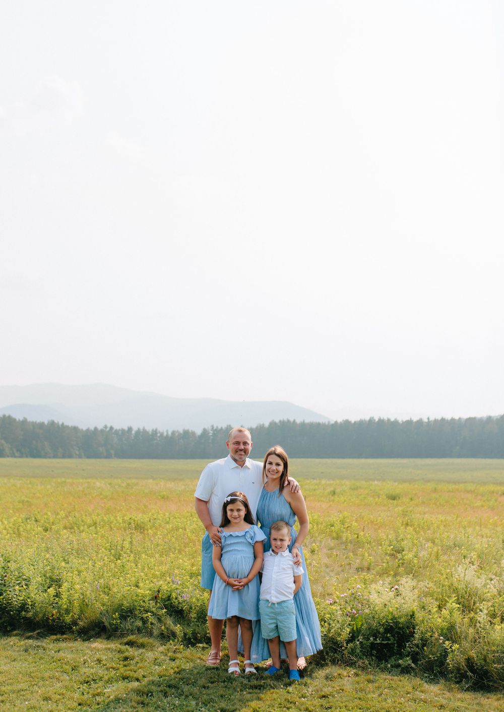 Family wearing coordinated light blue outfits poses together in a sunny meadow with forest backdrop.