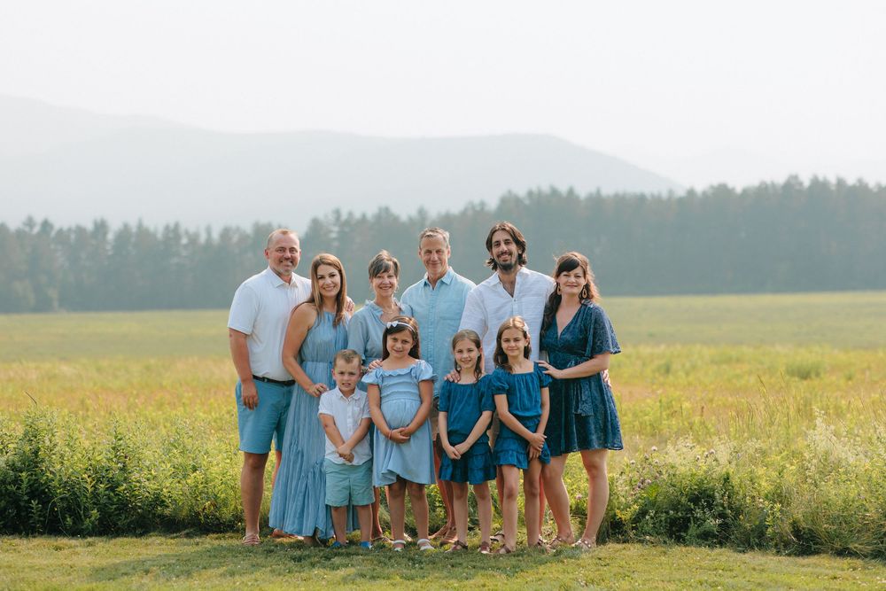 A group in coordinated blue and teal outfits poses together in a sunny meadow with forest in the background.