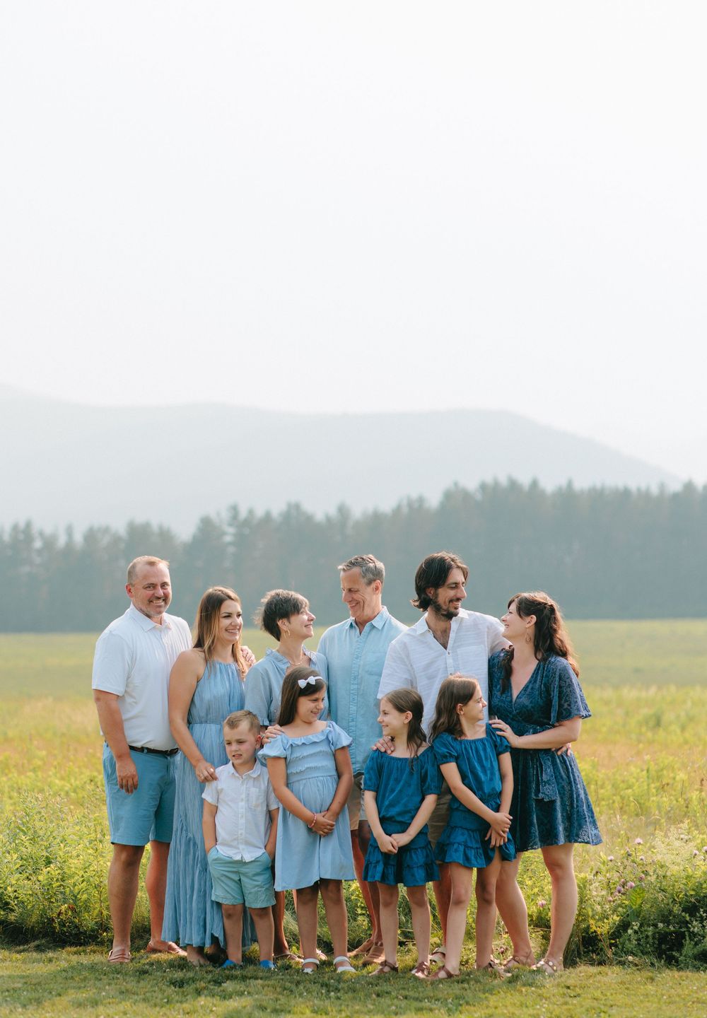 Group wearing coordinated blue outfits poses together in a sunny meadow with mountains in background.