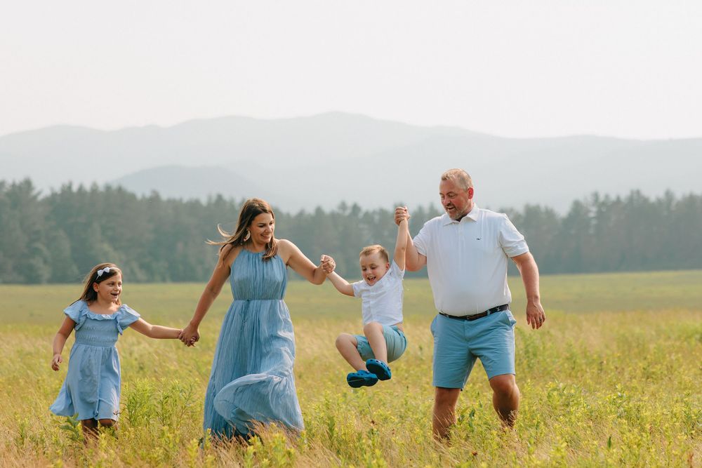 People holding hands while walking in a line through flowering meadow with forest backdrop.