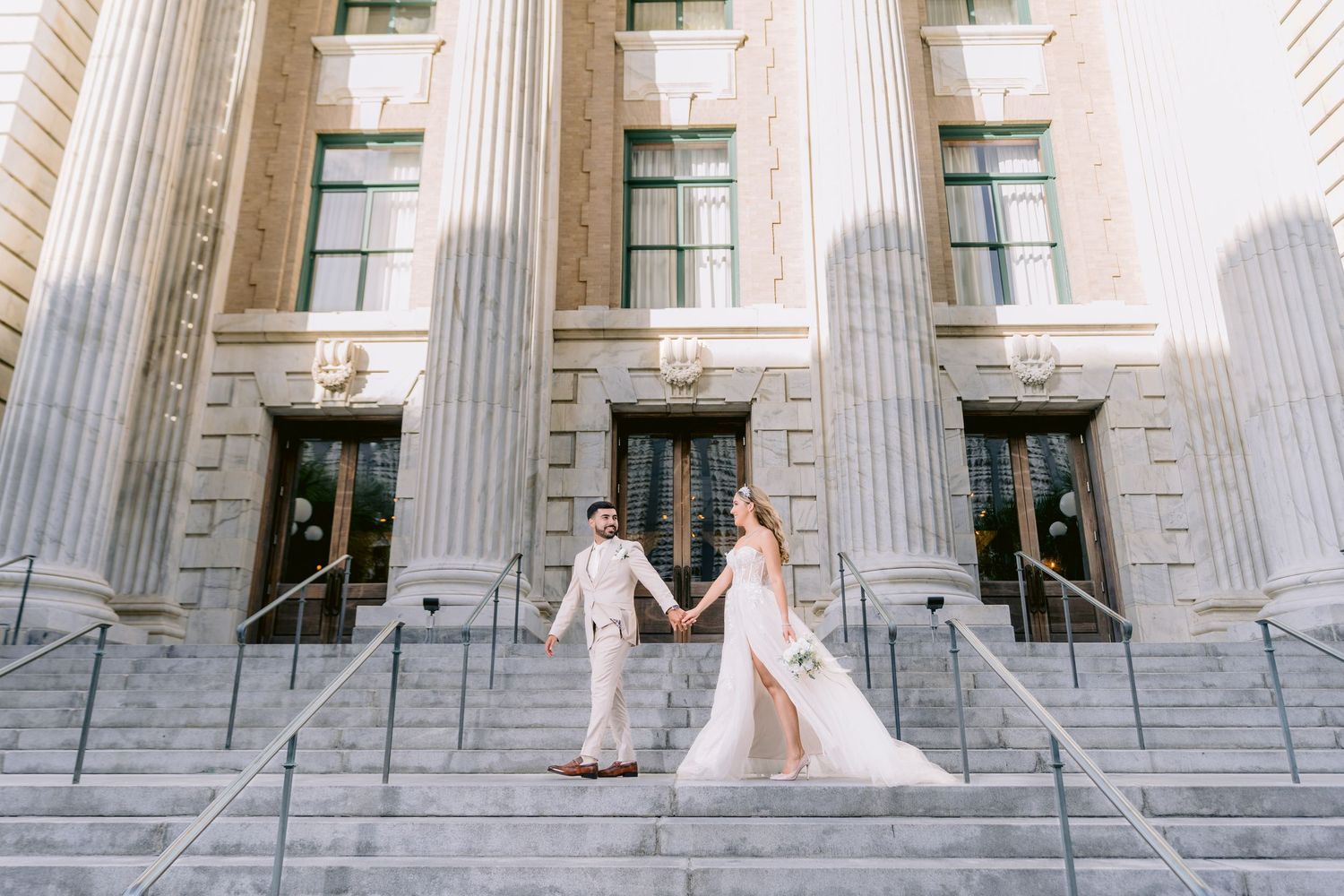 Elegant couple in wedding attire standing on grand marble steps of Le Meridien, Tampa,  building with neoclassical columns.