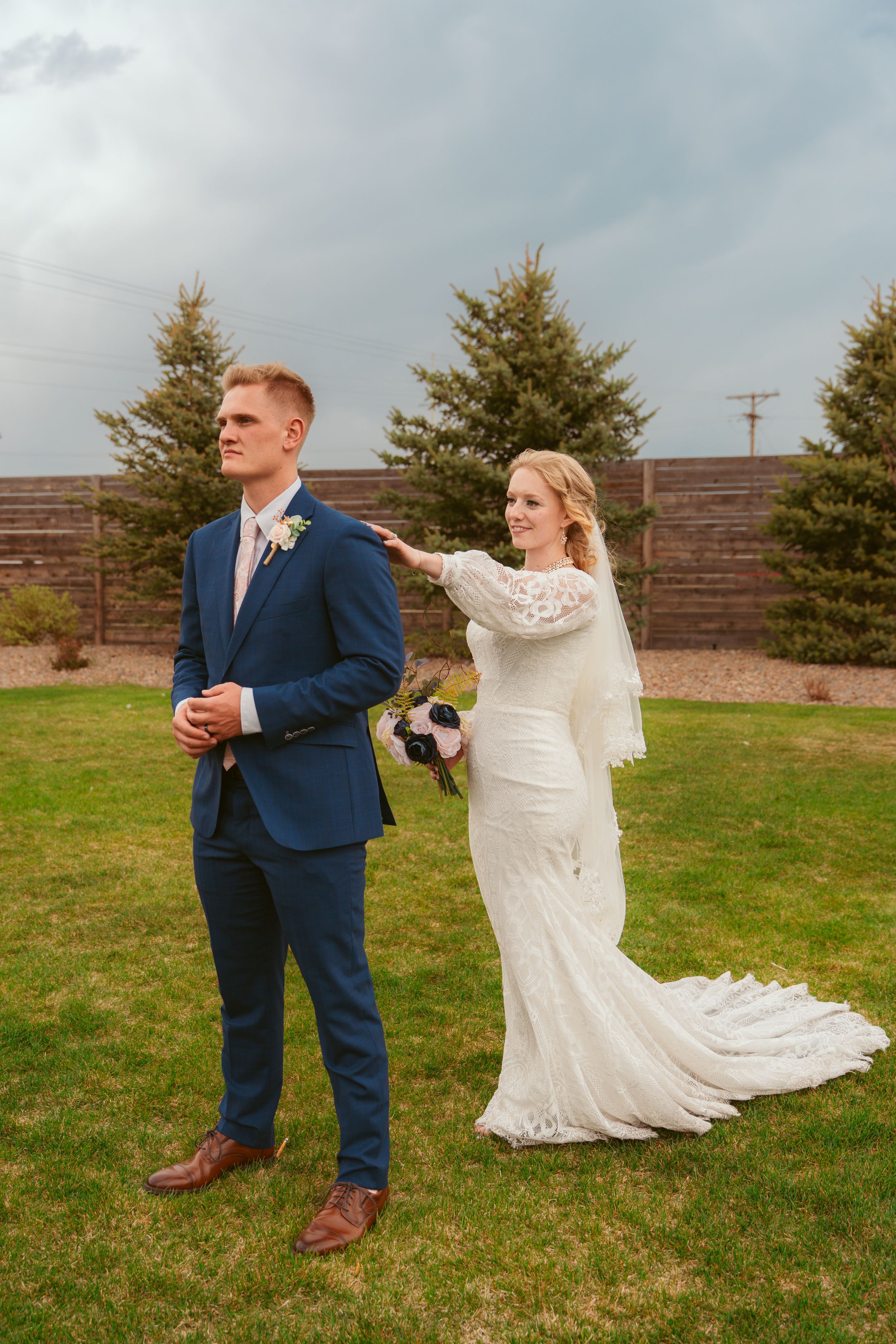 A couple shares their first look moment at an outdoor wedding venue with evergreen trees and brick wall backdrop.
