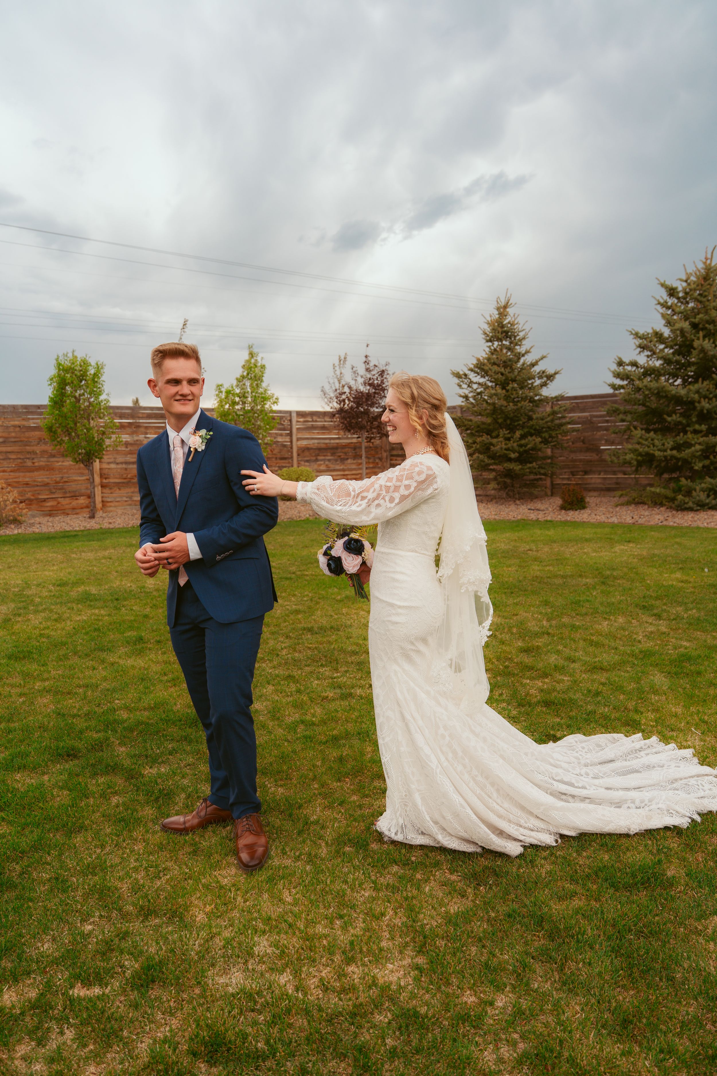 Wedding couple stands on a green lawn with evergreen trees and wooden fence in the backdrop on an overcast day.