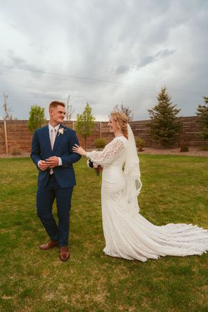 On a grassy field with evergreen trees, two figures embrace during a wedding photo shoot in long white gown and blue suit.