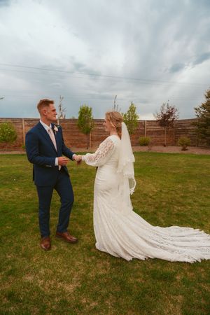 A newlywed couple stands together on a lawn with a wooden fence backdrop as the bride's lace dress trails elegantly behind her.