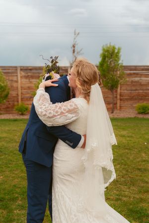 A couple embraces during their wedding ceremony in front of a wooden fence, with the bride wearing a lace gown and veil.