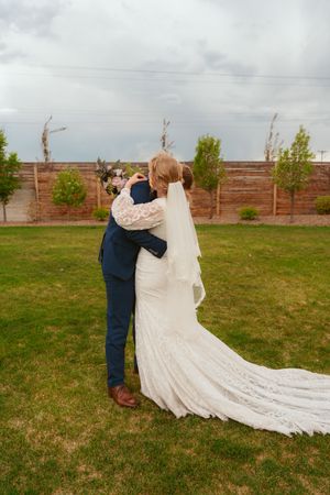 A romantic wedding couple embraces on a grassy lawn with a long flowing white lace train and brown fence in background.