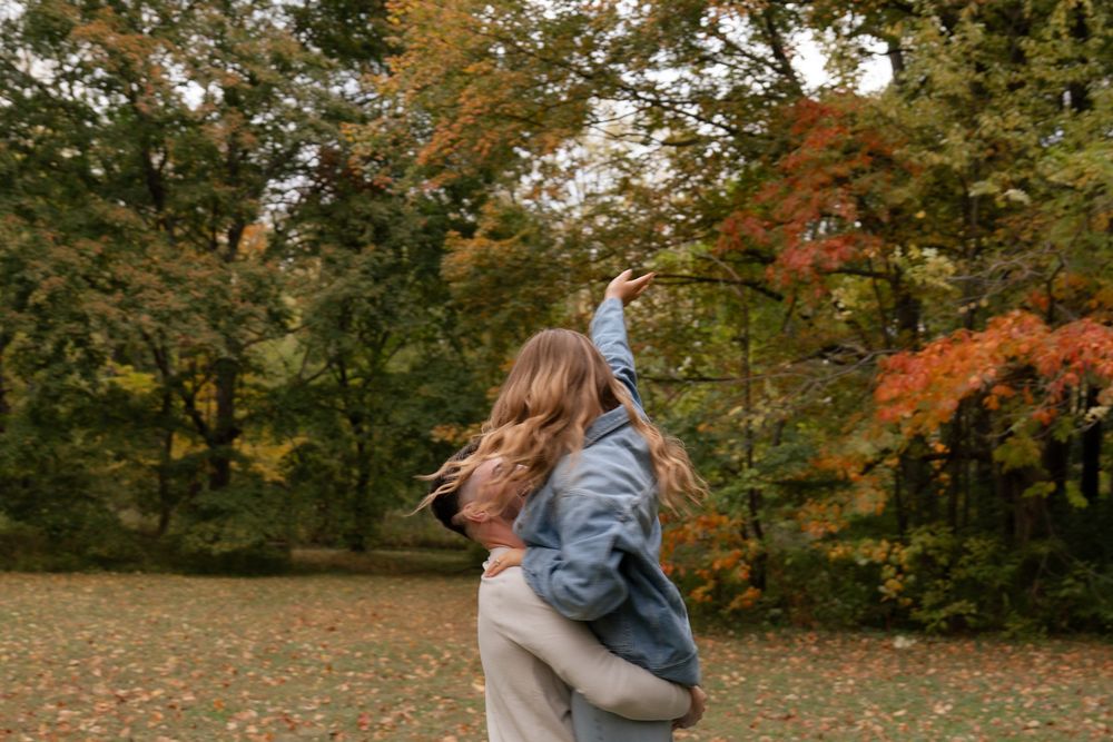 A couple embraces in a park surrounded by vibrant autumn foliage and falling leaves during a romantic photoshoot.