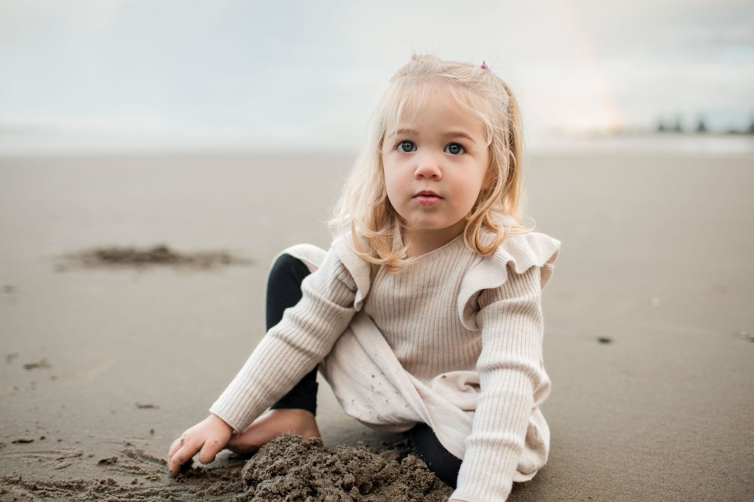 Child in beige sweater sitting on sandy beach during overcast day looks thoughtfully at the ground.