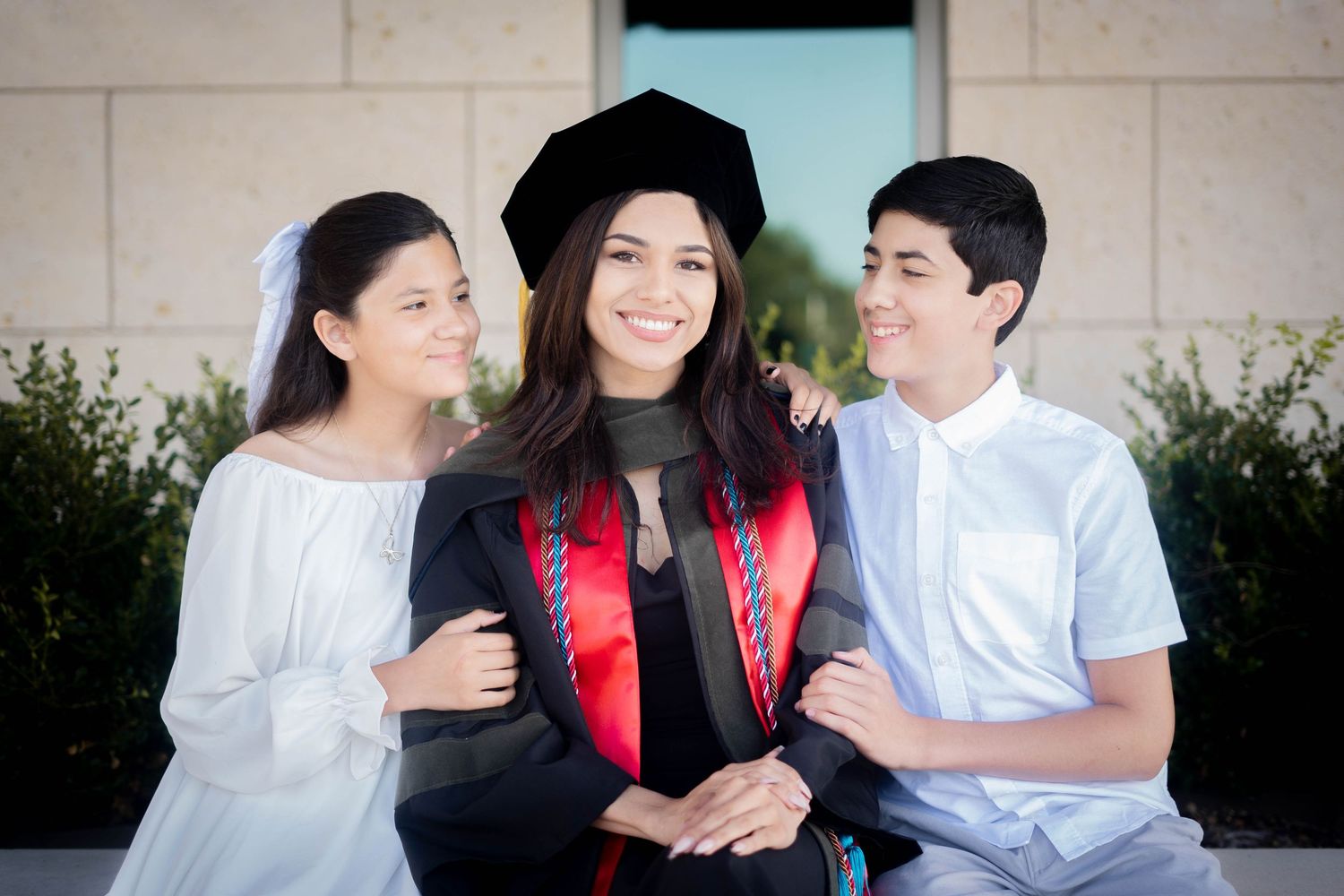 Smiling graduate in black cap and gown with red stole posing with two younger siblings.