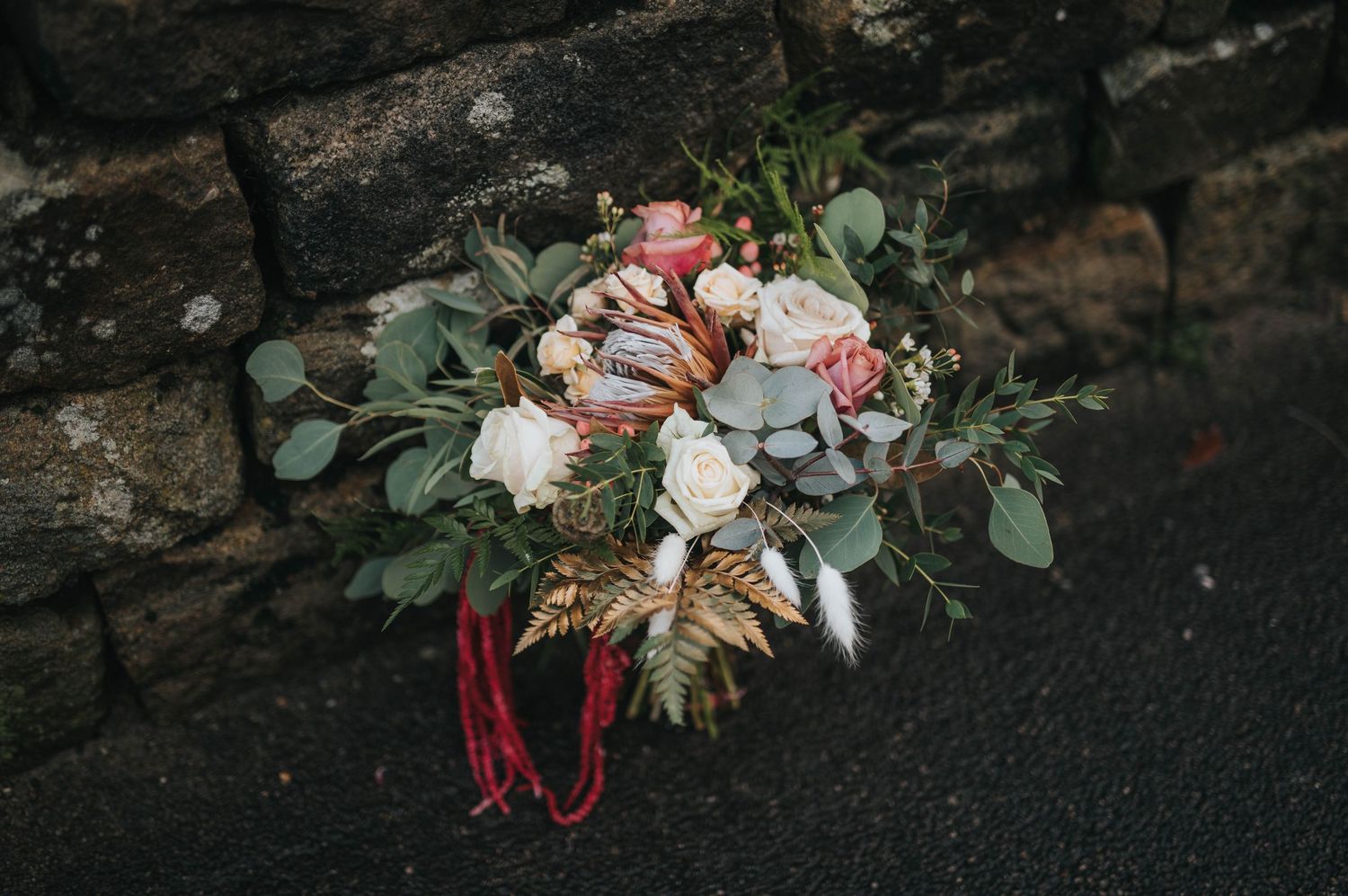 A rustic wedding bouquet with white and pink flowers, eucalyptus leaves, and burgundy ribbons rests against a stone wall.