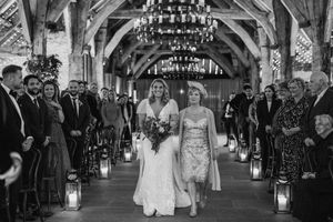 A wedding ceremony in an elegant barn venue with lantern-lined aisle and rustic wooden beam architecture.
