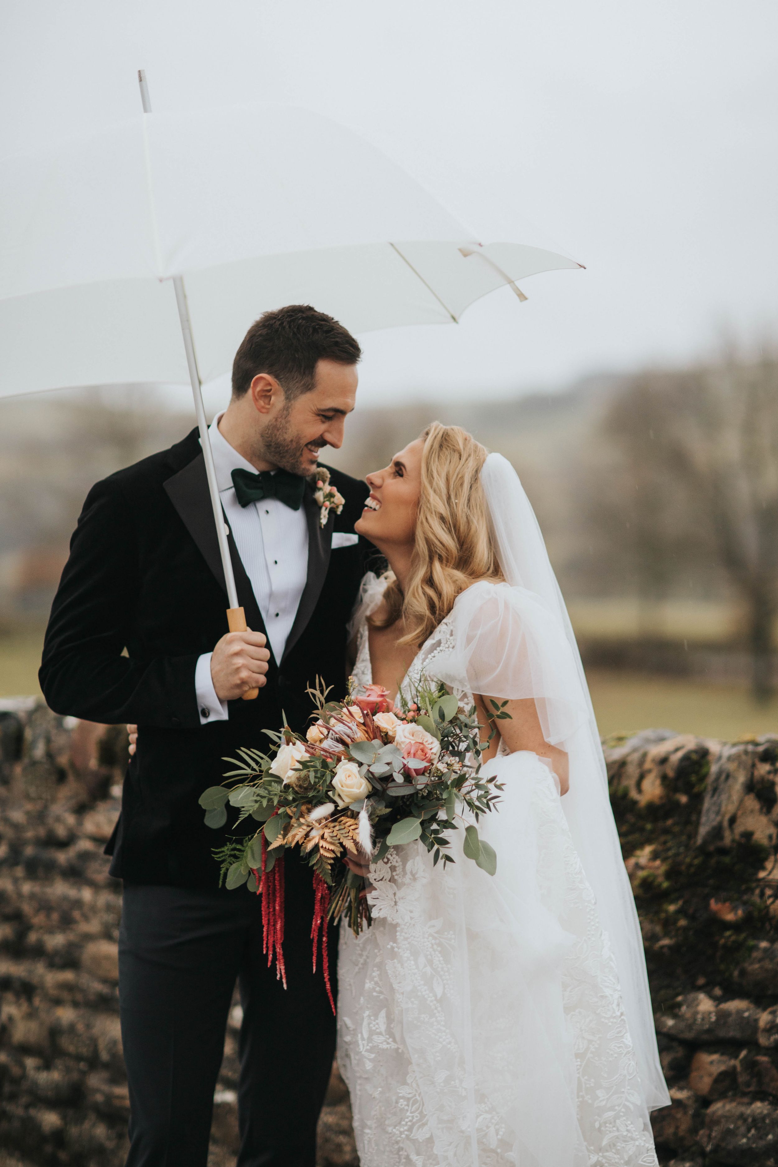 A bride and groom share an intimate moment under a white umbrella, with the bride holding a lovely pink and white bouquet.