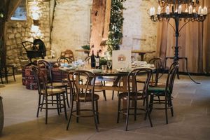 Rustic wooden dining set with chairs arranged around tables in stone-walled room with vintage chandelier lighting.