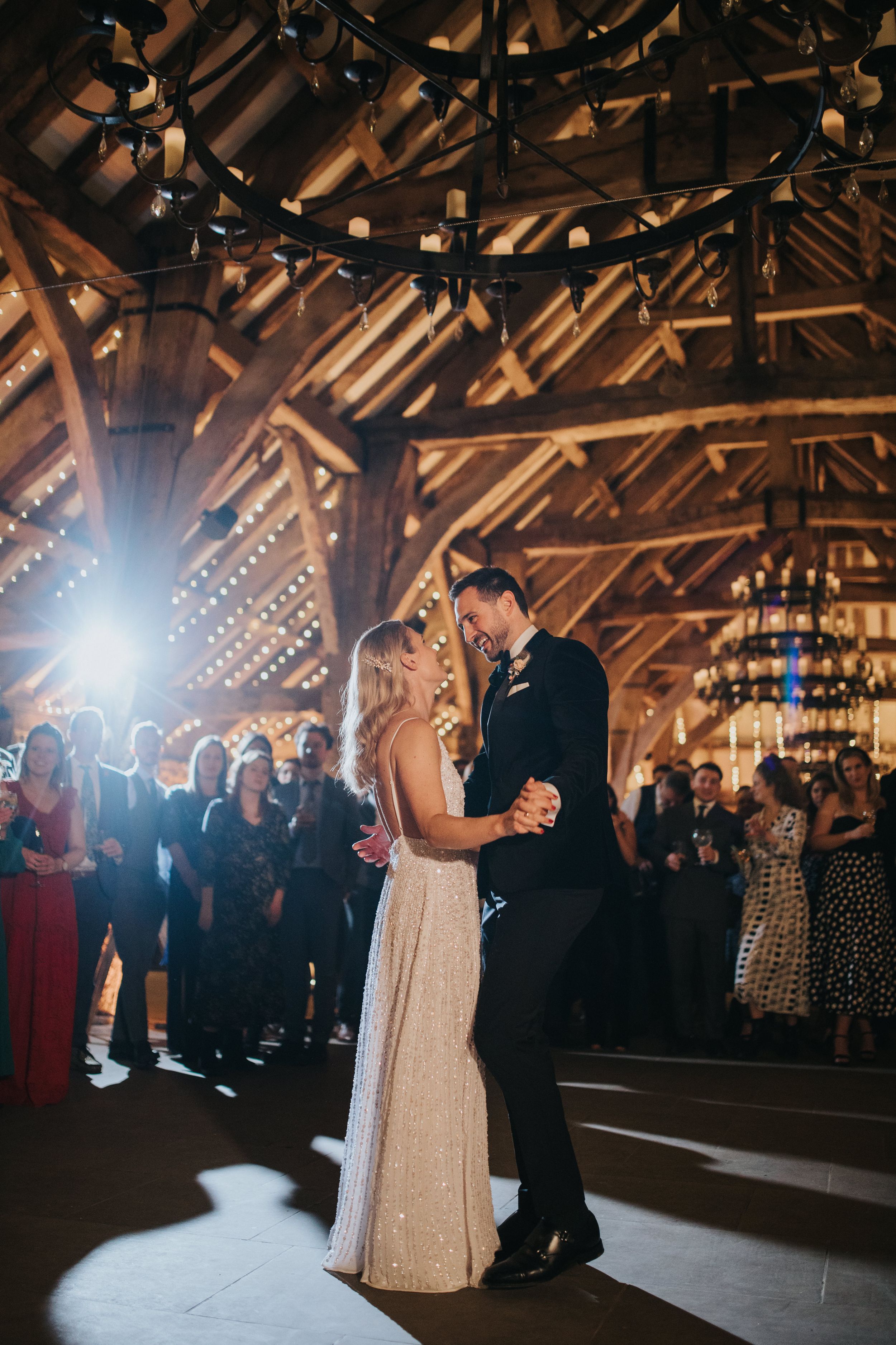 A couple shares their first dance at a rustic barn wedding reception with string lights overhead and guests watching.