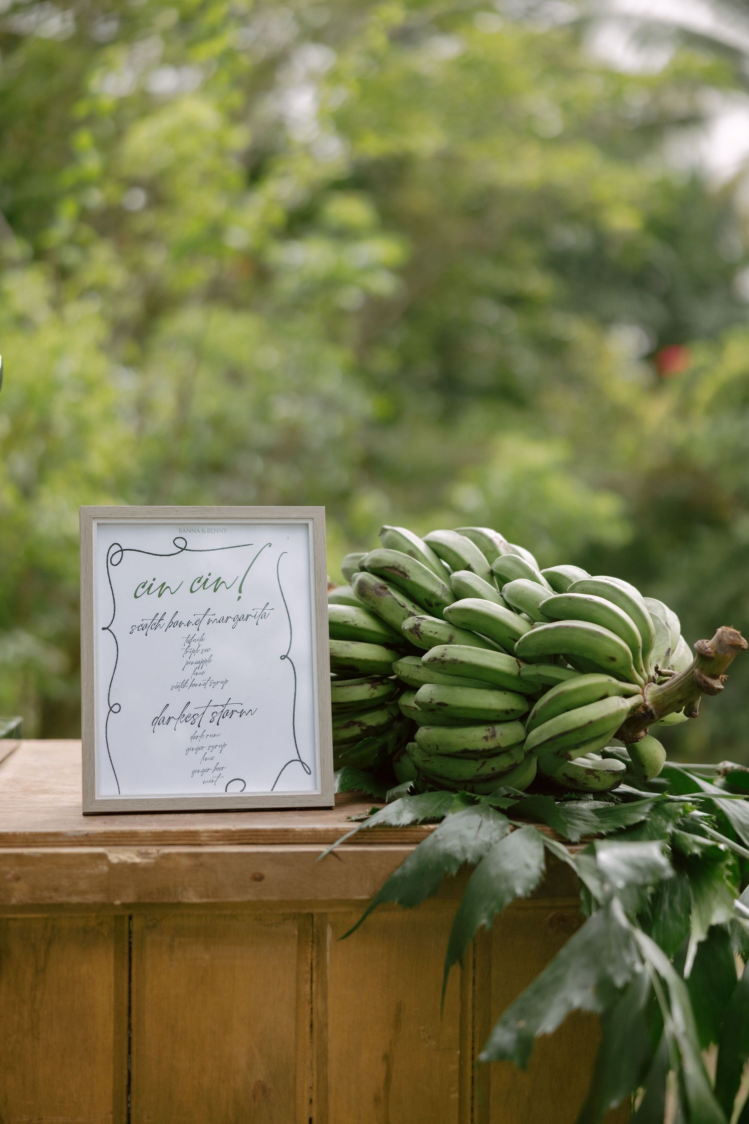 A bunch of green bananas sits next to a framed menu sign on a wooden surface in an outdoor garden setting.