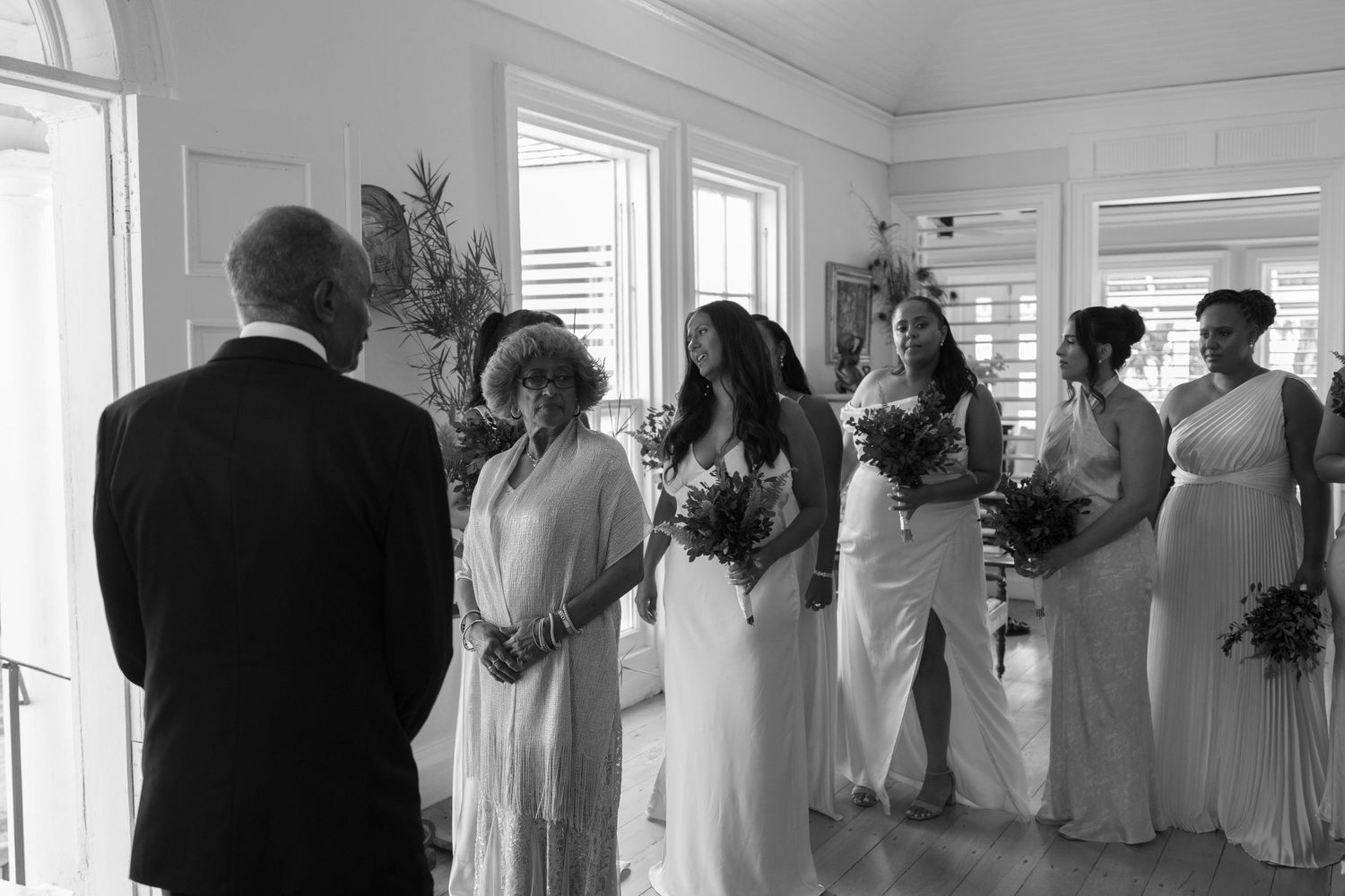 Black and white photo of a crowded reception room with people in formal white attire gathering near large windows.