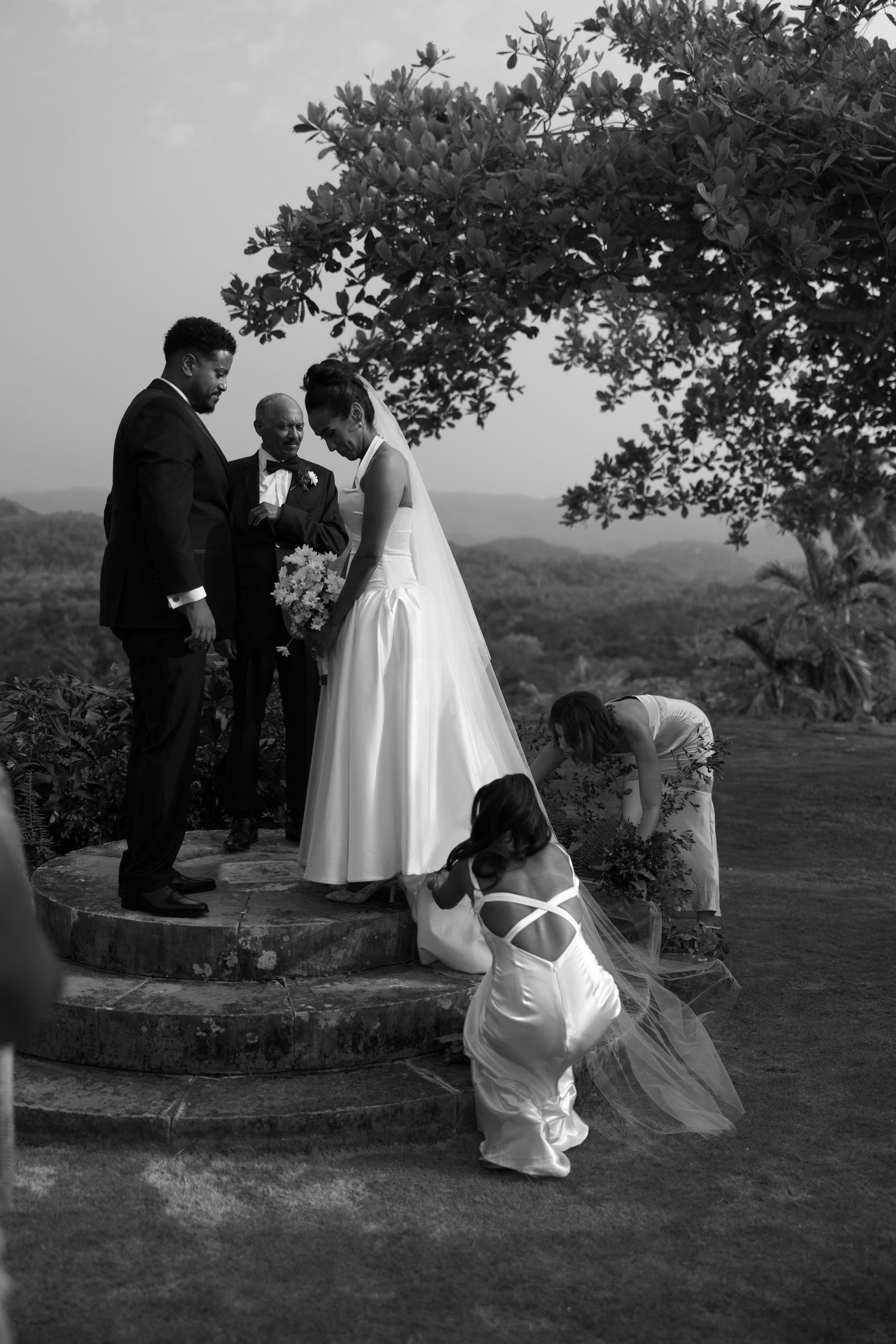 A black and white wedding ceremony photograph captured on a hilltop under a tree.