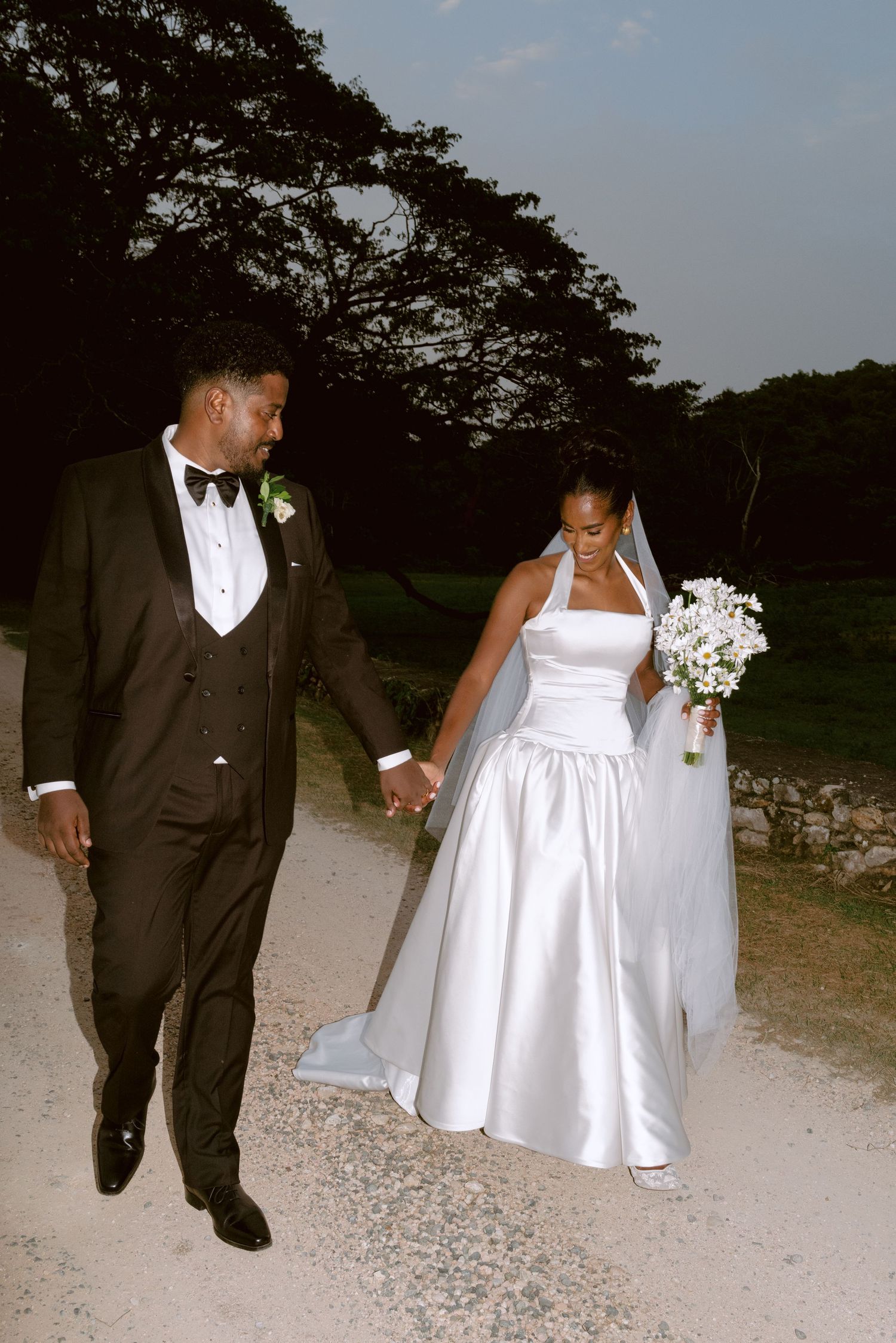 A bride and groom walk hand in hand along a gravel path, with the bride holding a white bouquet at dusk.