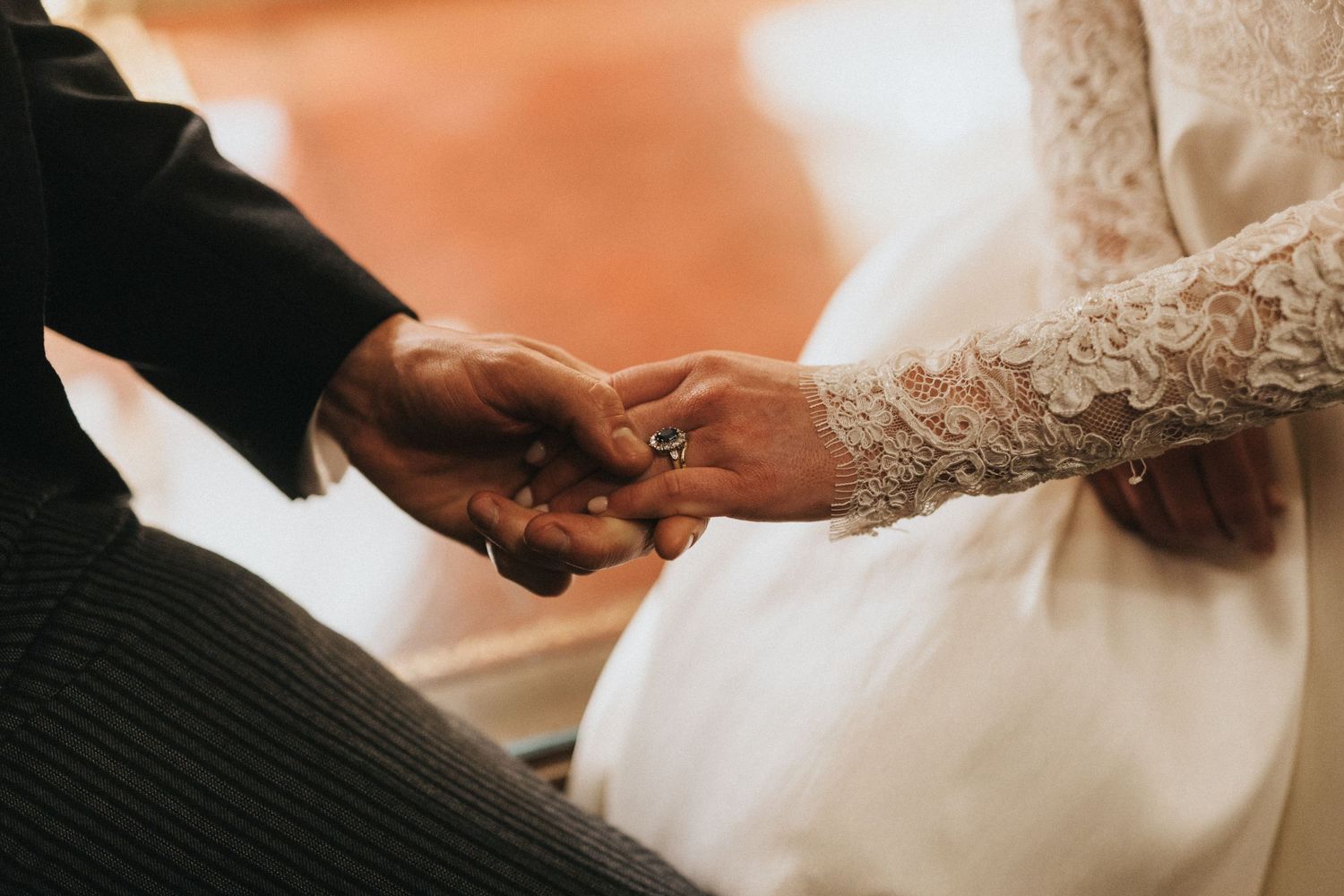 Close up shot of hands exchanging wedding rings during ceremony with lace sleeve detail and dark suit.