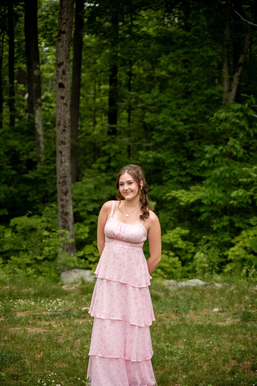 A person in a pink tiered formal dress poses in a lush green forest clearing.
